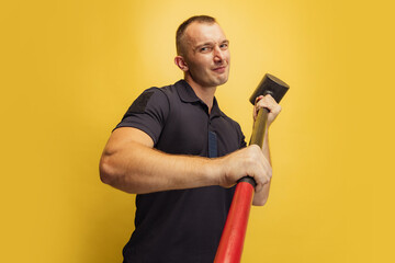 One young brutal man, firefighterposing with hammer over yellow studio background.
