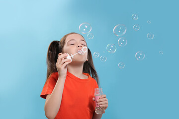 Little girl blowing soap bubbles on light blue background, space for text