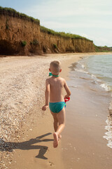 A boy runs along the beach in swimming trunks. He has a water pistol in his hands. Happy child on the sandy beach. Concept of happy life and relaxation by the ocean. Blond boy smiling at the camera