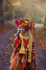 Herbstfee, ein kleines blondes Mädchen mit dem bunten Herbstkranz auf dem Kopf steht im bunten Herbstwald. Das Gesicht ist mit der Herbstmuster geschminkt. In der Hand ist eine Kastanie.