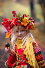 Herbstfee, ein kleines blondes Mädchen mit dem bunten Herbstkranz auf dem Kopf steht im bunten Herbstwald. Das Gesicht ist mit der Herbstmuster geschminkt.. In der Hand ist eine Kastanie.