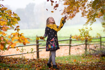 Blondes kleines Mädchen spielt im Wald mit den Herbstblättern, schmeißt sie in die Luft und schaut hoch. .Im Hintergrund ist ein Feld, Holzzaun, Nebel.