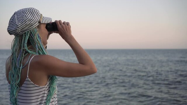 A young woman in a marine striped dress looks through binoculars at the sea or ocean