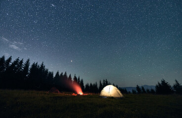 Fototapeta na wymiar Wide angle view on beautiful landscape in the mountains. Illuminated tourist tent and campfire under starry sky. Mountains range behind spruces. Concept of night camping and astrophotography