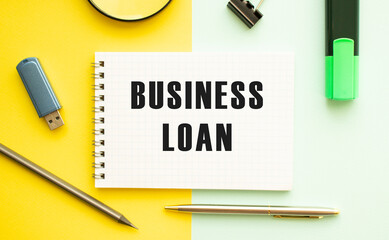 Notebook with text BUSINESS LOAN on office table with office supplies. Yellow color background.