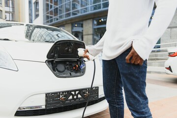 african man holding charge cable in on hand standing near luxury electric car.