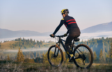 Male cyclist in cycling suit riding bike with coniferous trees and hills on background. Man bicyclist enjoying bicycle ride in mountains. Back view. Concept of sport, biking and active leisure.