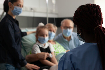 Selective focus of black asisstant discussing with caring family explaining healthcare treatment against clinical disease in hospital ward. Elderly senior woman resting in bed wearing face mask