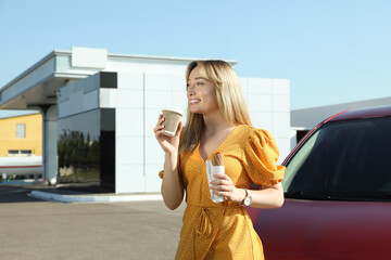 Beautiful young woman with hot dog drinking coffee near car at gas station
