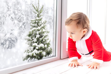 a baby boy in a Santa Claus costume looks out of the window in winter at a snow-covered Christmas tree, the concept of new year and Christmas