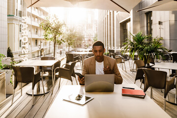 Young black man using cellphone while working with laptop at cafe