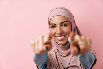 A smiling muslim woman wearing pink hijab pointing at the camera