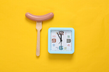 Time to eat. Sausage on fork and clock. Yellow background. Flat lay