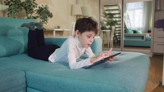 Curious Child Little school Boy Using Digital Tablet Technology Device Lying On Sofa Alone. Small Boy Hold Pad Computer Surfing Internet Play Game At Home. Children Tech Addiction Concept