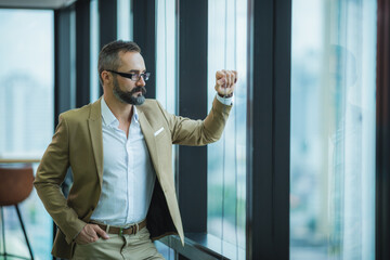 businessman look at the office's window, relax and chill moment during office time, modern lifestyle, smart business man in light brown outfits, caucasian man, city view background.