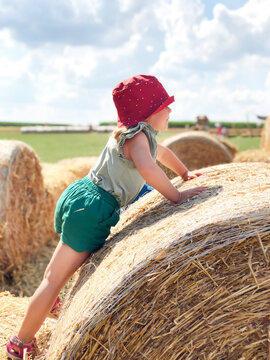 Little toddler girl having fun with running and jumping on hay stack or bale. Funny happy healthy child playing with straw. Active outdoors leisure with children on warm summer day. Kids and nature