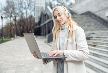 Young blonde woman in headphones holding laptop outdoors