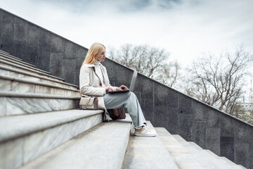 Young girl in trench coat is typing on laptop while sitting on the stairs. Remote work, freelance