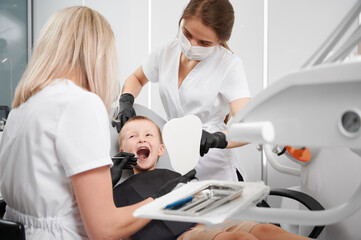 Adorable little boy sitting in dental chair while two female dentists checking kid teeth. Dentist...