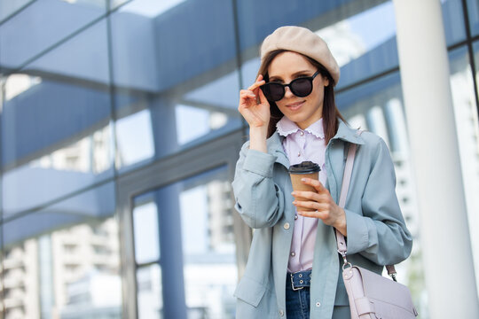 Stylish woman in sunglasses and beret holds a cup of coffee on the background of windows of the business center. Fashion, beauty, lifestyle