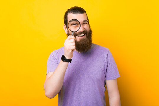 Cheerful bearded man is looking through a magnifying glass over yellow background.