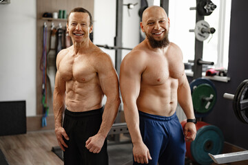 Two shirtless men standing in gym. Young man and his senior father exercise together in gym