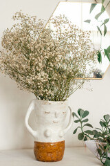 A bouquet of dried gypsophila flowers in a creative vase.A mirror in a decorative frame hangs on a white wall.Indoor plants.Home interior design.Biophillia design.Urban jungle.