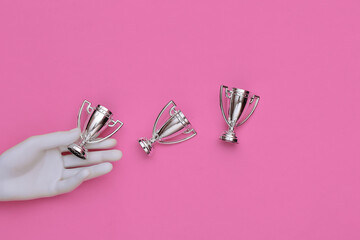 White mannequin hand holding championship cups on pink background. Top view