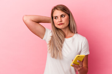 Young russian woman holding a mobile phone isolated on pink background touching back of head, thinking and making a choice.