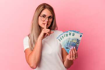 Young russian woman holding bills isolated on pink background keeping a secret or asking for silence.