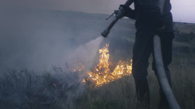 A firefighter holds a hose with water in his hands, extinguishes a burning flame. Nature is on fire: grass, steppe, forests and fields. Water from hose is sprayed onto the fire. Close-up, slow motion