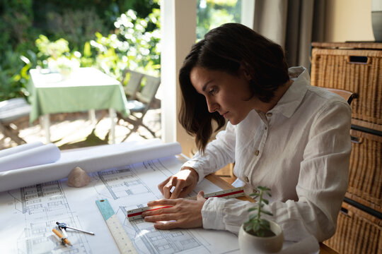 Caucasian female architect in living room, sitting at table working, drawing plans