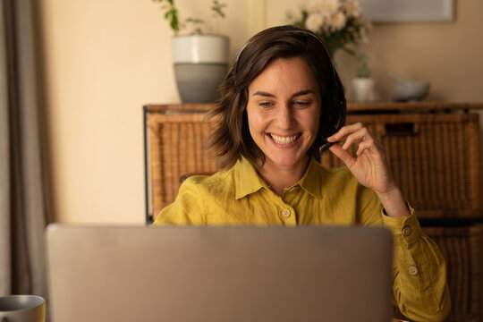 Smiling caucasian woman in living room, sitting at table working, using laptop wearing headset