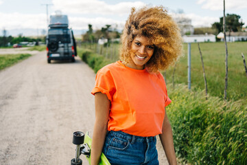 Cheerful black woman with longboard walking in countryside