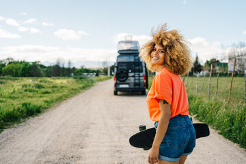 Cheerful black woman with longboard walking in countryside