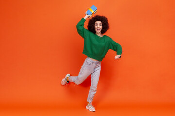 Fototapeta na wymiar Full length portrait of happy woman with curly hair wearing green casual style sweater throwing blue wrapped present box, has excited expression. Indoor studio shot isolated on orange background.