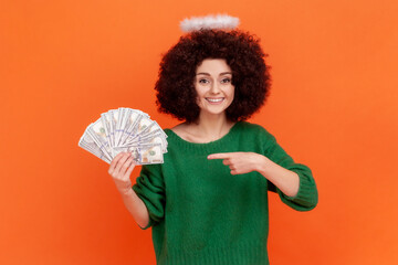 Young adult angelic woman with curly hair wearing green casual style sweater with nimb over head holding and pointing at fan of dollars banknotes. Indoor studio shot isolated on orange background.
