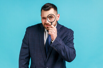 Young adult handsome businessman wearing dark official style suit holding magnifying glass and...