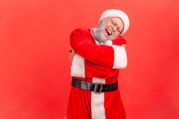 Fototapeta na wymiar Elderly man with gray beard wearing santa claus costume hugging himself, feeling comfortable, narcissistic egoistic person, happy expression. Indoor studio shot isolated on red background.