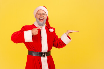Fototapeta na wymiar Extremely happy elderly man with gray beard wearing santa claus costume presenting copy space on his palm, looking at camera with open mouth. Indoor studio shot isolated on yellow background.