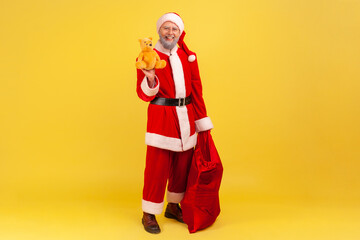 Fototapeta na wymiar Full length of elderly man with gray beard wearing santa claus costume holding red big bag with present, giving cute bear as a present, Indoor studio shot isolated on yellow background.