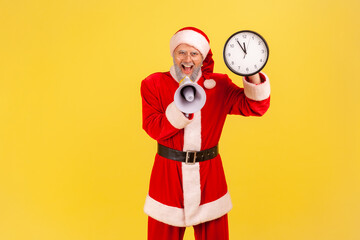 Portrait of yelling excited elderly man with gray beard wearing santa claus costume screaming in megaphone and showing wall clock in hands. Indoor studio shot isolated on yellow background.