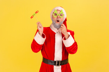 Fototapeta na wymiar Excited elderly man with gray beard wearing santa claus costume holding paper cards on sticks, celebrating christmas and congratulating with new year. Indoor studio shot isolated on yellow background.