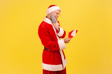 Fototapeta na wymiar Profile portrait of elderly man with gray beard wearing santa claus costume being touched with sensitive letter, reading Christmas congratulations. Indoor studio shot isolated on yellow background.