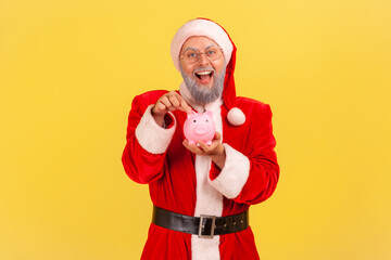 Fototapeta na wymiar Excited elderly man with gray beard wearing santa claus costume standing with open mouth, with positive expression, showing piggy bank to camera. Indoor studio shot isolated on yellow background.