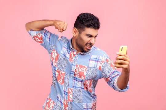 Portrait of angry mad man with beard wearing blue casual style shirt, having unpleasant video call, clenched fist to hit mobile phone, crazy expression. Indoor studio shirt isolated on pink background