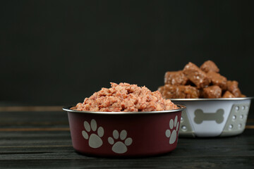 Different pet food in feeding bowls on wooden table against dark background, space for text