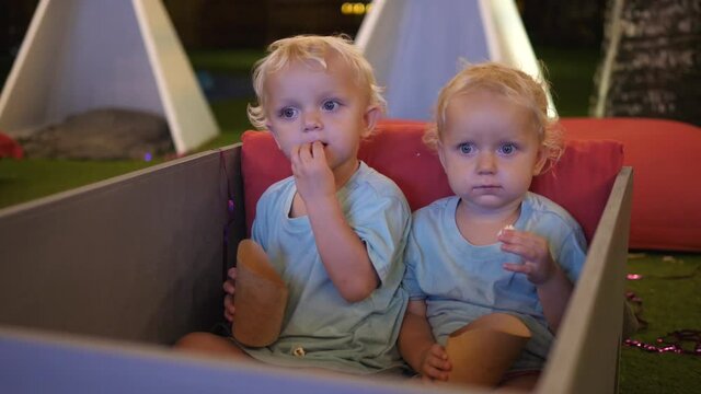Two cute white twins watching movie with curiosity and eating kids popcorn