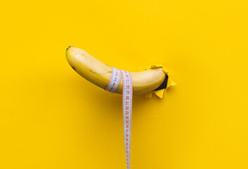 A banana with a tape measure wrapped around it appears through a torn hole in yellow paper. The concept of healthy food, diet and potency. Copy space. - obrazy, fototapety, plakaty