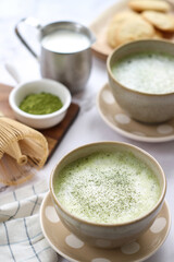 Bowls of healthy organic matcha green tea with mousse and chasen whisk with its powder, pot of milk and cookie in background.  Matcha  is an powerful source benefits for health and diet people   